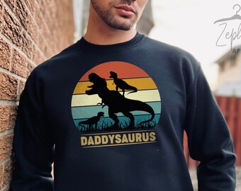 May include: A black sweatshirt featuring a vintage-style graphic of a dinosaur family. The graphic depicts a large dinosaur with two smaller dinosaurs on its back, all silhouetted against a sunset. The text "DADDYSAURUS" is printed below the graphic.