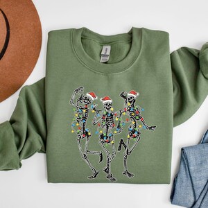 May include: A green sweatshirt with a festive design of three skeletons wearing Santa hats and Christmas lights. The skeletons are dancing and smiling.