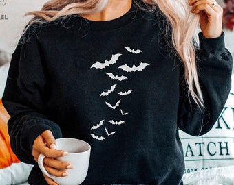 Könnte beinhalten: Schwarzes Sweatshirt mit einer weißen Silhouette von Fledermäusen, die diagonal über die Vorderseite fliegen. Das Sweatshirt wird von einer Person getragen, die auf einem Sofa sitzt.