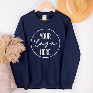 Peut inclure: Un sweat-shirt bleu marine avec le texte "YOUR logo HERE" dans un cercle blanc. Le sweat-shirt est suspendu à un cintre en bois. Un chapeau de paille et un vase avec des plantes séchées sont également dans l'image.