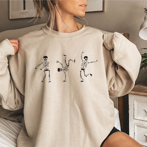 May include: A beige sweatshirt with a graphic of three skeletons dancing. The skeletons are black and the sweatshirt is a light beige color.
