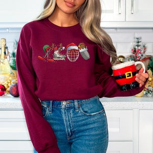 May include: A maroon sweatshirt with a Christmas-themed hockey design. The design features a hockey stick, skates, a hockey helmet, and a Santa hat.