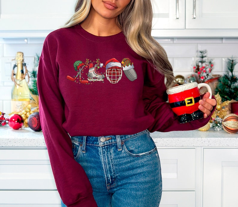 May include: A maroon sweatshirt with a Christmas-themed hockey design. The design features a hockey stick, skates, a hockey helmet, and a Santa hat.