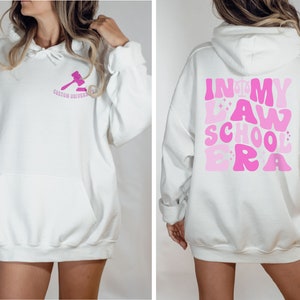 May include: White hoodie with a pink gavel graphic on the front and the text "In My Law School Era" in pink on the back.