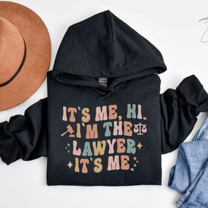 Peut inclure: Sweat-shirt &agrave; capuche noir avec le texte "It's me, hi. I'm the lawyer. It's me." en police r&eacute;tro color&eacute;e. Le texte est entour&eacute; de petites &eacute;toiles.