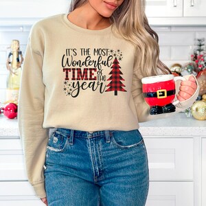 Puede incluir: Una sudadera beige con el texto "It's the most Wonderful Time of the year" y un gr&aacute;fico de &aacute;rbol de Navidad de cuadros rojos y negros.