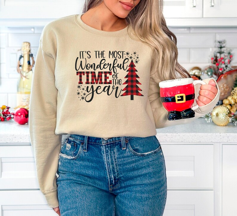 Puede incluir: Una sudadera beige con el texto "It's the most Wonderful Time of the year" y un gr&aacute;fico de &aacute;rbol de Navidad de cuadros rojos y negros.