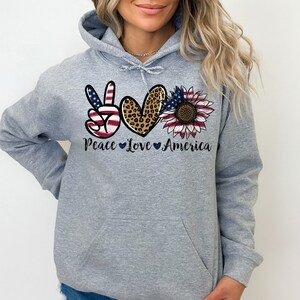May include: Gray hooded sweatshirt with a peace sign, heart, and sunflower design. The peace sign is an American flag, the heart is leopard print, and the sunflower is red, white, and blue. The text "Peace Love America" is printed below the design.
