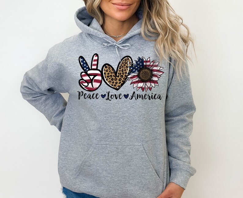 May include: Gray hooded sweatshirt with a peace sign, heart, and sunflower design. The peace sign is an American flag, the heart is leopard print, and the sunflower is red, white, and blue. The text "Peace Love America" is printed below the design.
