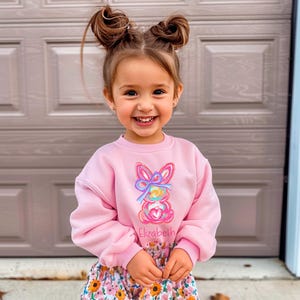 May include: A pink sweatshirt with a colorful bunny graphic and the name "Elizabeth" printed on the front. The sweatshirt has long sleeves and a crew neck.
