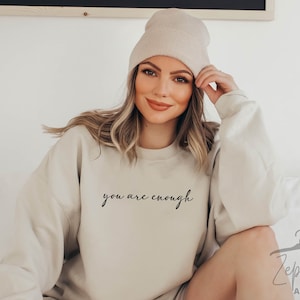 May include: A beige sweatshirt with the text "you are enough" in black script. The model is wearing a beige beanie and has long blonde hair.