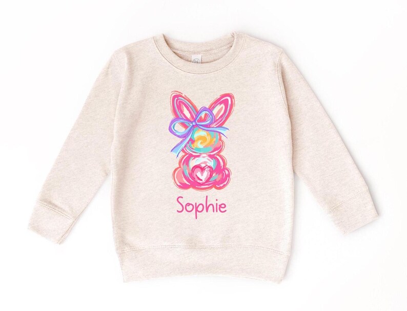 May include: A beige long-sleeved sweatshirt with a colorful bunny rabbit graphic and the name "Sophie" printed below it.