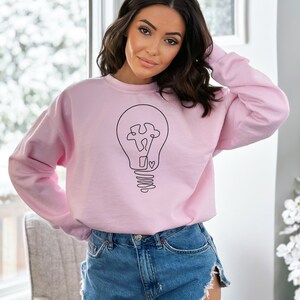 May include: A pink sweatshirt with a black line drawing of a lightbulb with a puzzle piece inside and a heart at the bottom.