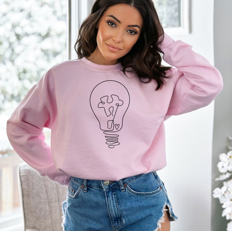 May include: A pink sweatshirt with a black line drawing of a lightbulb with a puzzle piece inside and a heart at the bottom.