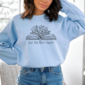 Peut inclure: Sweat-shirt bleu clair avec un contour noir d'un livre ouvert avec des fleurs qui en sortent. Le texte "Just One More Chapter" est imprim&eacute; sous le livre.