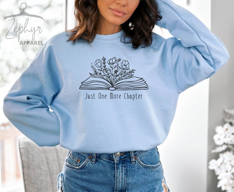 Peut inclure: Sweat-shirt bleu clair avec un contour noir d'un livre ouvert avec des fleurs qui en sortent. Le texte "Just One More Chapter" est imprim&eacute; sous le livre.