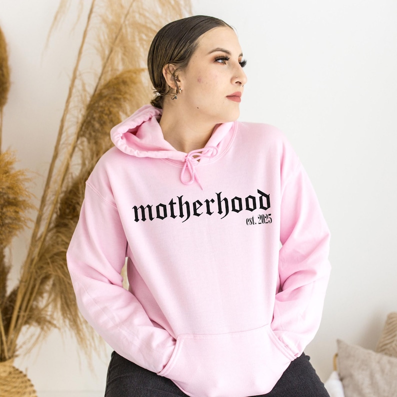 Op de afbeelding: Een roze hoodie met een zwart gotisch lettertype ontwerp dat "motherhood est. 2025" zegt.