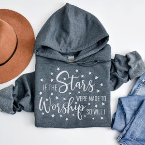 May include: A gray hooded sweatshirt with the text "If the Stars Were Made to Worship So Will I" in white, surrounded by stars. A brown hat and blue jeans are also in the image.
