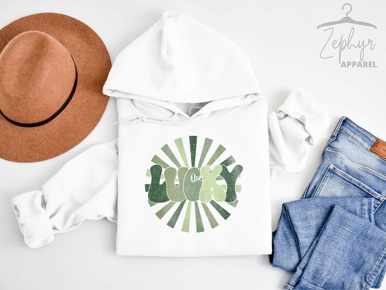 Peut inclure: Sweat-shirt &agrave; capuche blanc avec un motif vert et marron du mot "LUCKY" dans un style r&eacute;tro avec un design en forme de soleil.