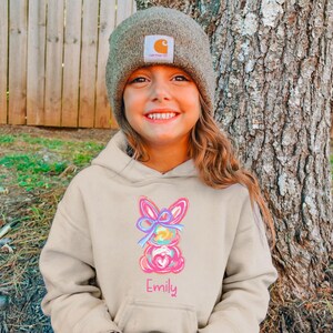 May include: A beige hoodie with a colorful Easter bunny design and the name "Emily" embroidered on the front. The hoodie is worn by a young person wearing a brown beanie.
