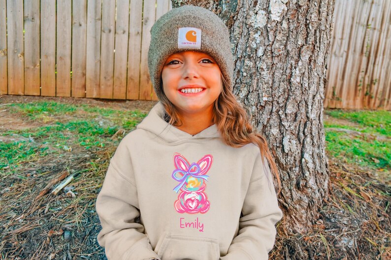 May include: A beige hoodie with a colorful Easter bunny design and the name "Emily" embroidered on the front. The hoodie is worn by a young person wearing a brown beanie.