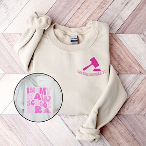 May include: A beige sweatshirt with a pink gavel and the text "Custom University" on the front. A separate image shows a white sweatshirt with the text "In My Law School Era" in pink.