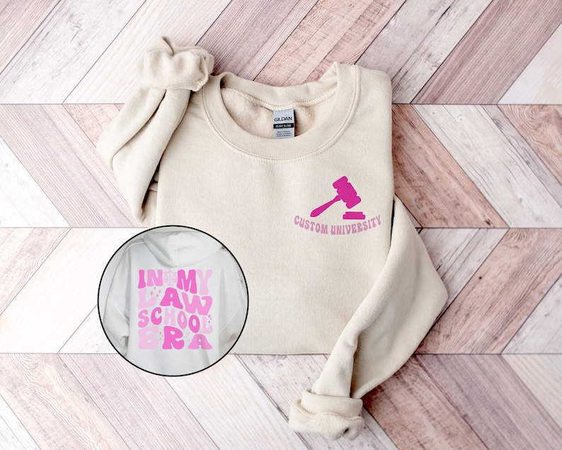 May include: A beige sweatshirt with a pink gavel and the text "Custom University" on the front. A separate image shows a white sweatshirt with the text "In My Law School Era" in pink.