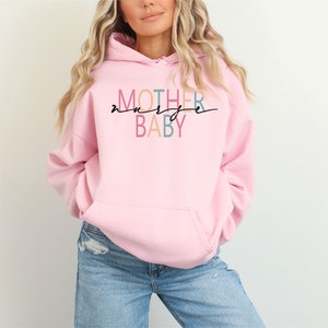Puede incluir: Sudadera rosa con el texto "MOTHER" en colores arco&iacute;ris y "BABY" en azul y rosa. El texto est&aacute; escrito en una fuente cursiva.