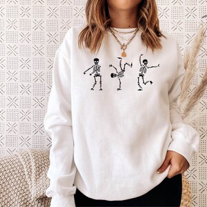 May include: White sweatshirt with a graphic of three skeletons dancing. The skeletons are black and white and are in different poses. The sweatshirt is a crew neck and has long sleeves.