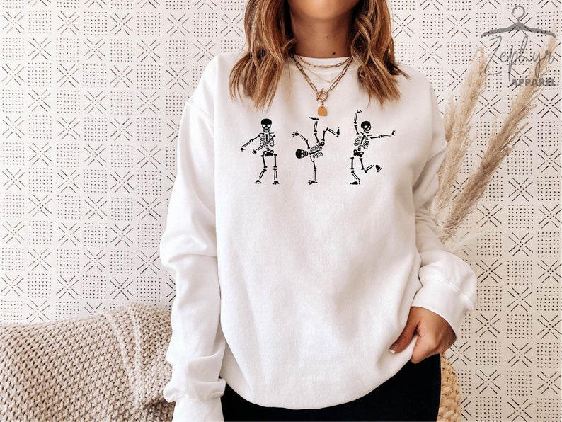 May include: White sweatshirt with a graphic of three skeletons dancing. The skeletons are black and white and are in different poses. The sweatshirt is a crew neck and has long sleeves.