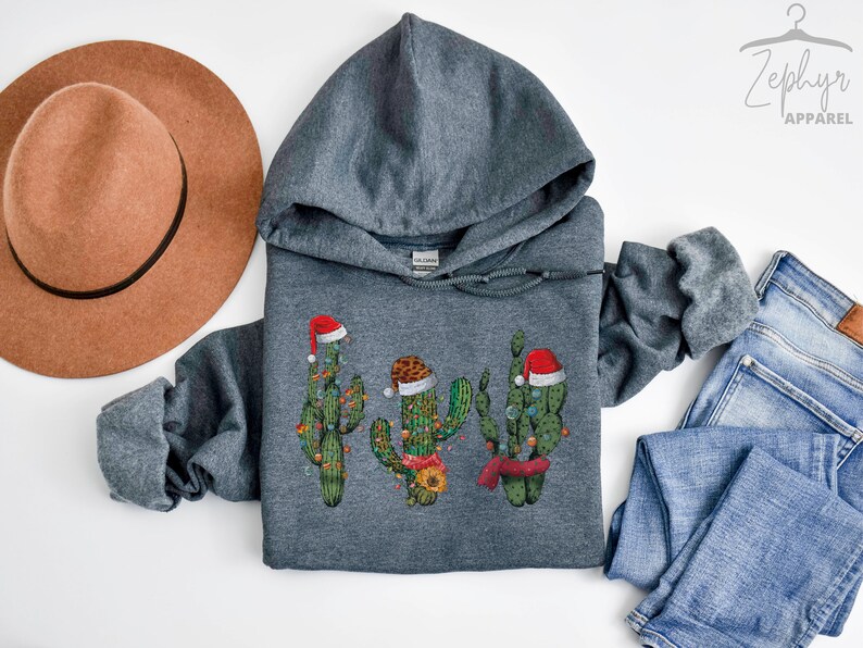 May include: A dark gray hooded sweatshirt with a Christmas cactus design. The cactus is wearing a Santa hat and has Christmas lights on it. The sweatshirt is on a white background.