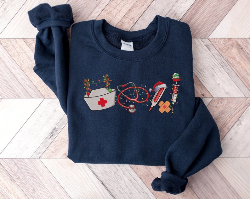 Puede incluir: Una sudadera azul marino con un dise&ntilde;o navide&ntilde;o que incluye un gorro de enfermera, un estetoscopio, un term&oacute;metro, una venda y una jeringa. El dise&ntilde;o est&aacute; formado por varios s&iacute;mbolos m&eacute;dicos decorados con luces de Navidad y un gorro de Pap&aacute; Noel.