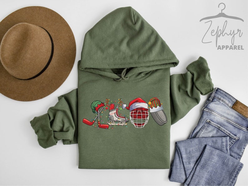May include: A green hooded sweatshirt with a Christmas-themed hockey design. The design features a hockey stick, a skate, a hockey helmet, and a puck, all decorated with Christmas lights and Santa hats.