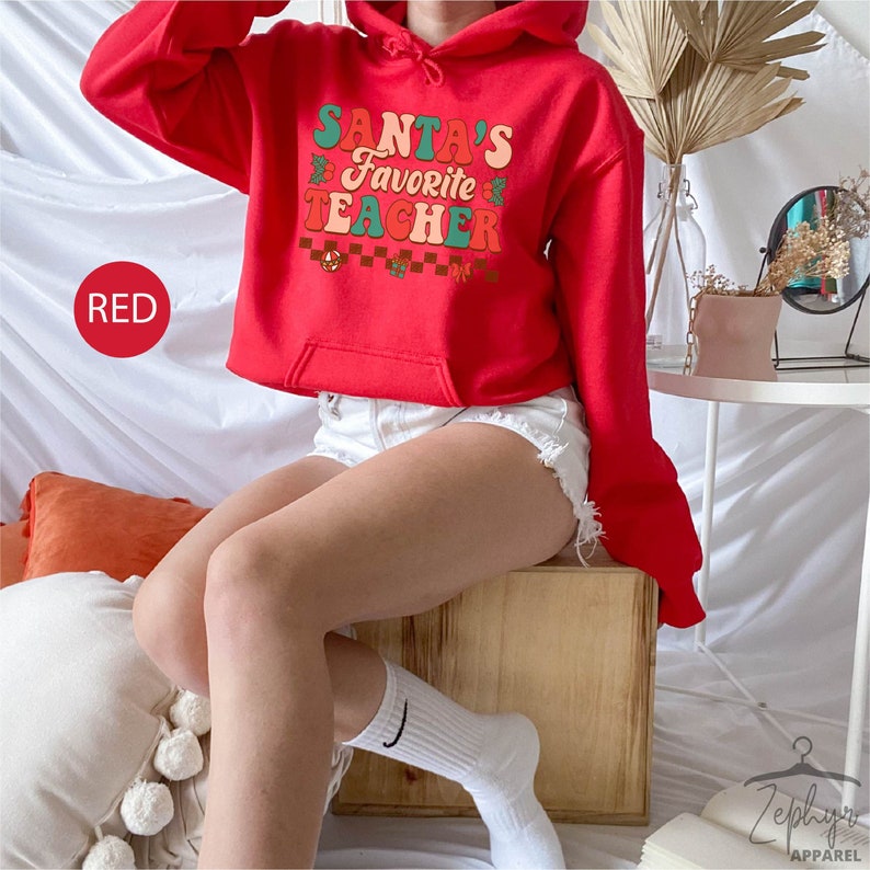 K&ouml;nnte beinhalten: Ein roter, kurzer Hoodie mit dem Text "Santa's Favorite Teacher" in einer Retro-Schrift mit gr&uuml;nen und orangen Akzenten. Das Model tr&auml;gt wei&szlig;e Jeans-Shorts und wei&szlig;e Socken.