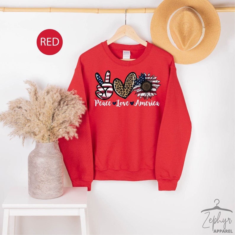 May include: Red sweatshirt with a peace sign, heart, and sunflower graphic. The graphic features an American flag, leopard print, and sunflower. The text "Peace Love America" is printed below the graphic.