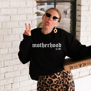 Op de afbeelding: Een vrouw draagt een zwart sweatshirt met de tekst "motherhood est. 2025" in witte gotische letters. Ze draagt een zonnebril en een gouden zonnebloem oorring. Ze houdt een pizzadoos in haar rechterhand en geeft een peace-teken met haar linkerhand.