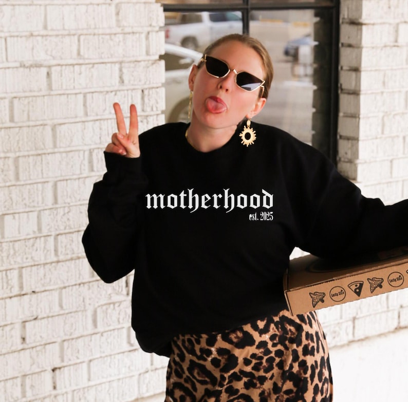 Op de afbeelding: Een vrouw draagt een zwart sweatshirt met de tekst "motherhood est. 2025" in witte gotische letters. Ze draagt een zonnebril en een gouden zonnebloem oorring. Ze houdt een pizzadoos in haar rechterhand en geeft een peace-teken met haar linkerhand.