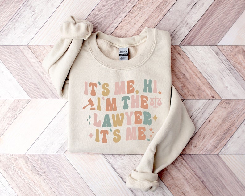 Peut inclure: Un sweat-shirt beige avec le texte "It's me, hi. I'm the lawyer. It's me." en lettres r&eacute;tro color&eacute;es. Un marteau et une balance de la justice sont inclus dans le design.