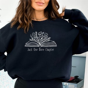 Peut inclure: Sweat-shirt noir avec un graphique blanc d'un livre ouvert avec des fleurs qui en sortent. Le texte "Just One More Chapter" est sous le livre.