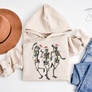 May include: A beige hoodie with a graphic of three skeletons wearing Santa hats and Christmas lights. The hoodie has a drawstring hood and a kangaroo pocket.