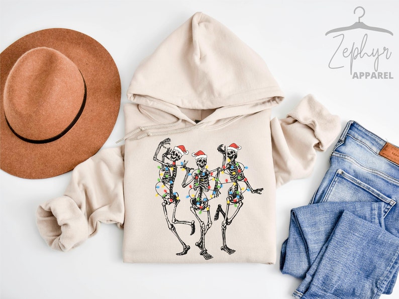 May include: A beige hoodie with a graphic of three skeletons wearing Santa hats and Christmas lights. The hoodie has a drawstring hood and a kangaroo pocket.