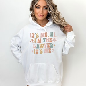 Peut inclure: Sweat-shirt &agrave; capuche blanc avec le texte "It's me, hi. I'm the lawyer. It's me." dans une police r&eacute;tro avec un graphique de marteau et de balance.