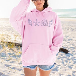 May include: A light pink hoodie with a front pocket and a drawstring hood. The hoodie features a design of blue seashell illustrations. The background shows a beach with sand and ocean.