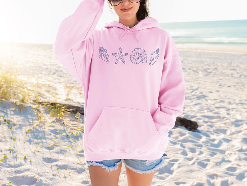 May include: A light pink hoodie with a front pocket and a drawstring hood. The hoodie features a design of blue seashell illustrations. The background shows a beach with sand and ocean.