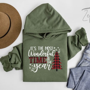 Puede incluir: Sudadera con capucha verde con el texto "It's the most Wonderful Time of the Year" en blanco con un gr&aacute;fico de &aacute;rbol de Navidad de cuadros rojos y negros.