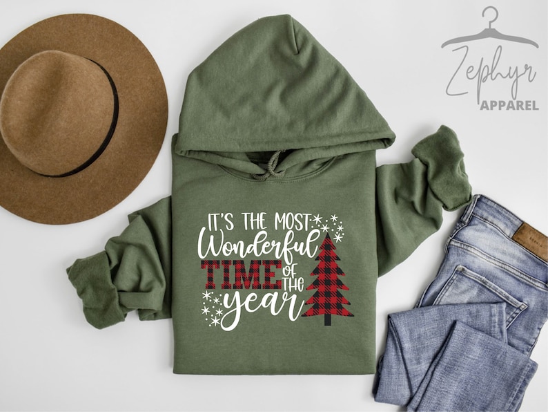 Puede incluir: Sudadera con capucha verde con el texto "It's the most Wonderful Time of the Year" en blanco con un gr&aacute;fico de &aacute;rbol de Navidad de cuadros rojos y negros.