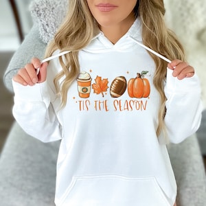 May include: White hoodie with a graphic of a coffee cup, a leaf, a football, and a pumpkin. The text "'Tis the season" is printed below the graphic.
