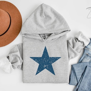 May include: A grey hoodie with a blue distressed star graphic. The hoodie has a white hood lining and is shown with a brown hat and blue jeans.