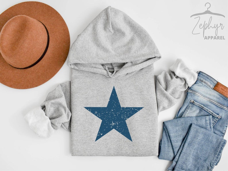 May include: A grey hoodie with a blue distressed star graphic. The hoodie has a white hood lining and is shown with a brown hat and blue jeans.