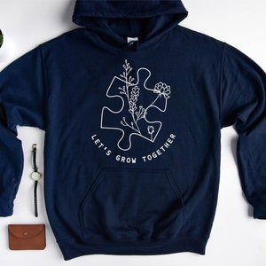 May include: Navy blue hoodie with a white outline of a puzzle piece with flowers and the text "Let's Grow Together".